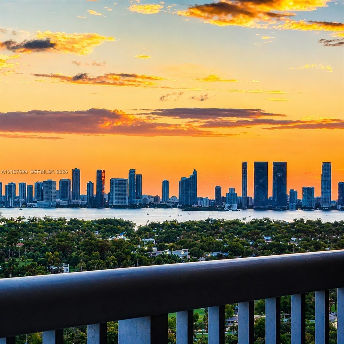 Elevated balcony with a metal railing overlooking a cityscape, waterfront, and lush tree canopy under a vibrant sunset sky