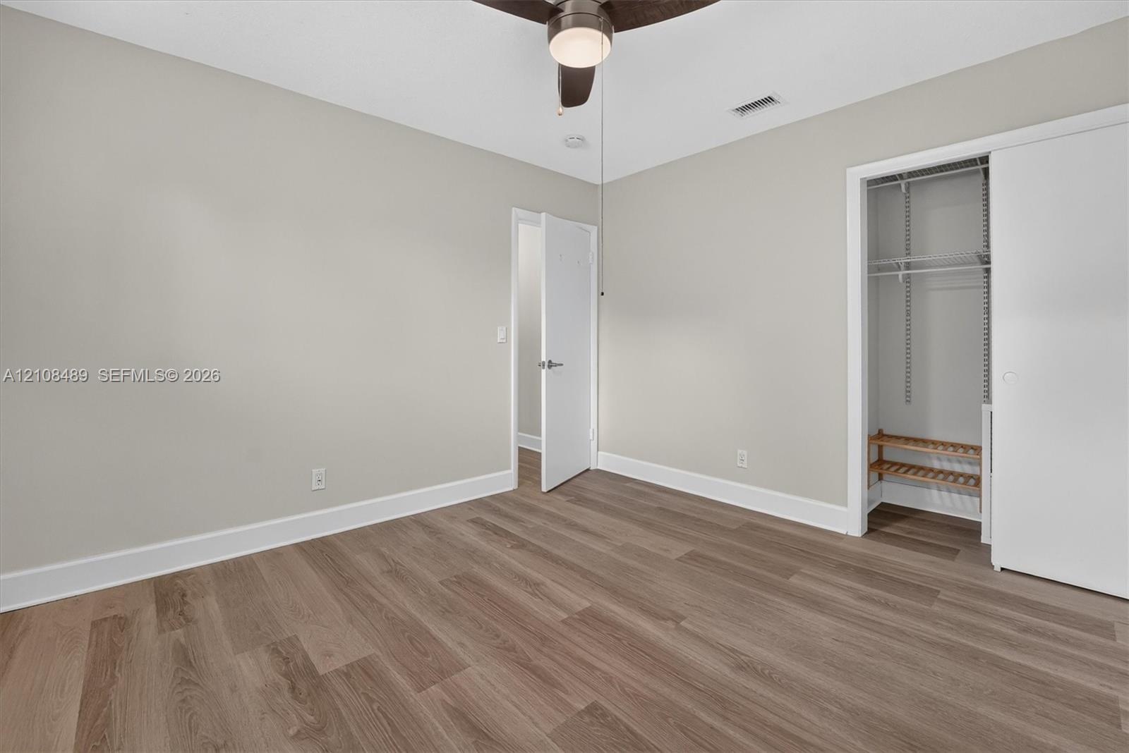 Neutral-toned interior space featuring wood-finish flooring, a ceiling fan with integrated lighting, and a closet with built-in wire shelving