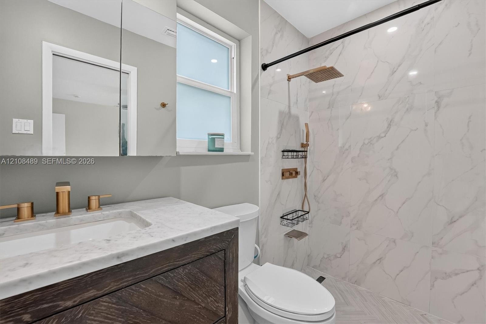 Bathroom featuring a vanity with wood-finish cabinetry, a light-toned stone countertop, brass-finish fixtures, a shower enclosure with large-format st