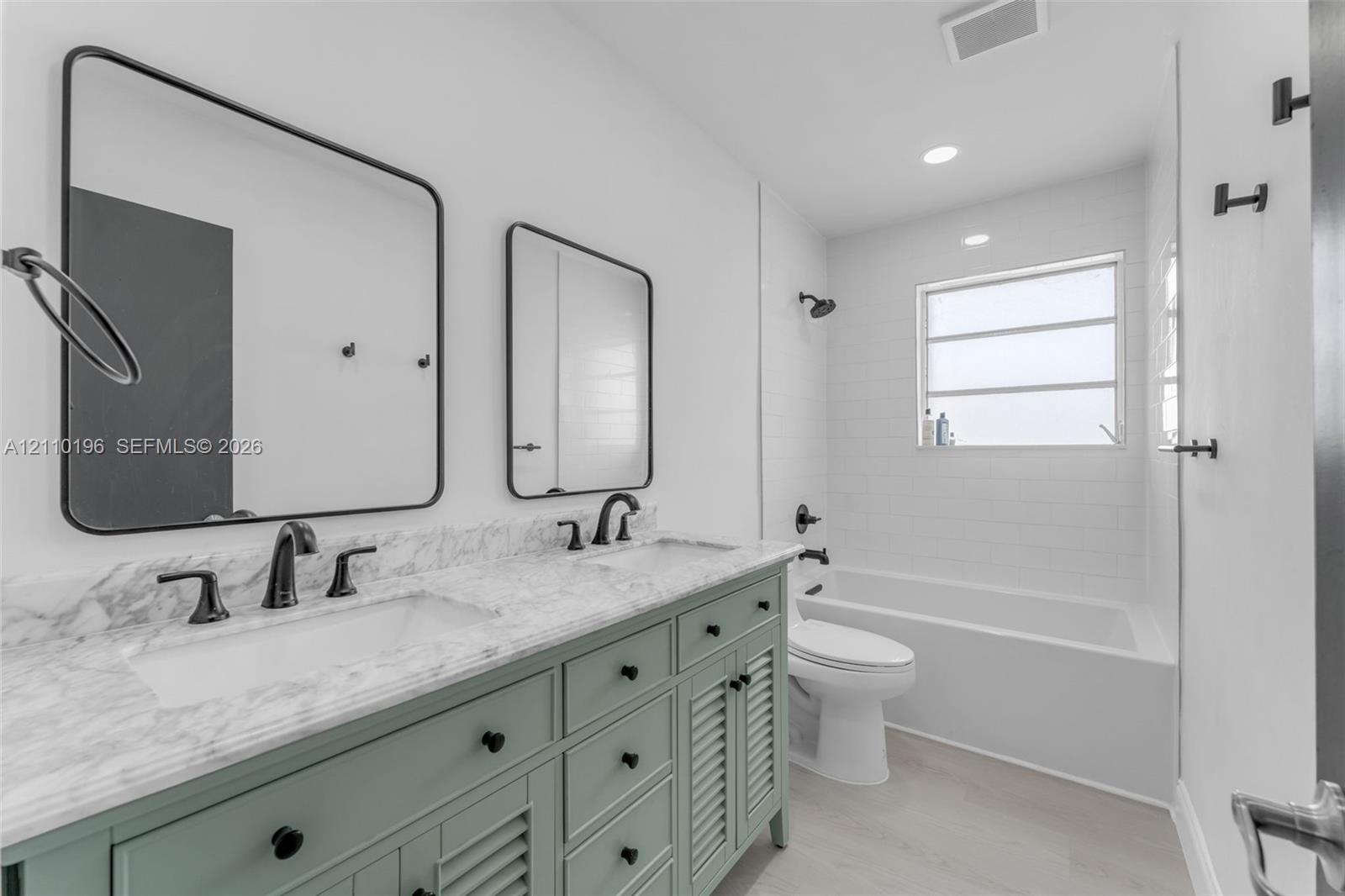 Double vanity with stone-look countertop, green cabinetry with louvered door detailing, matte black fixtures, rectangular wall-mounted mirrors, and a 