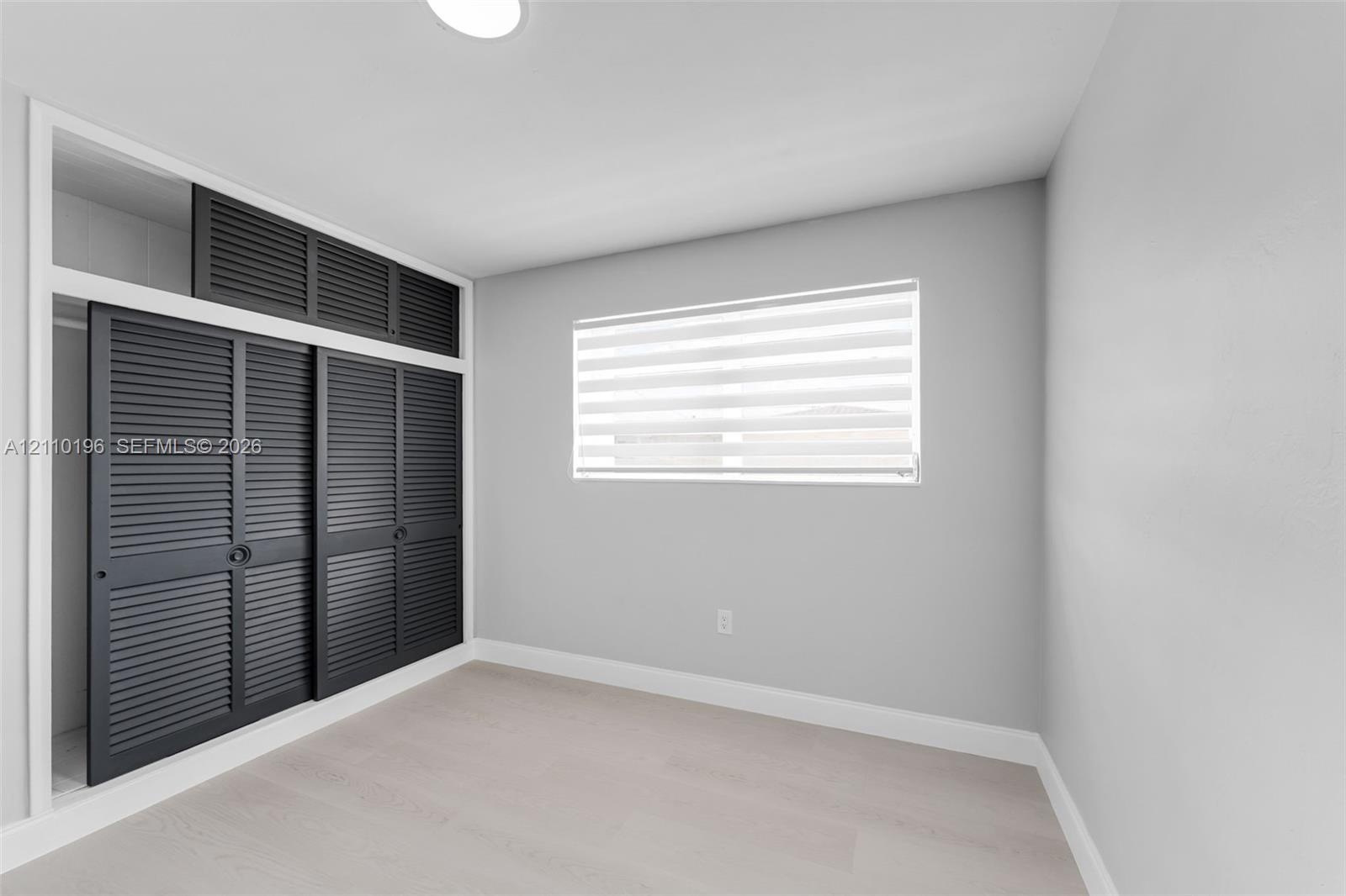 Interior space featuring light-toned wood-finish flooring, charcoal-colored louvered sliding closet doors with built-in overhead storage, and dual-lay