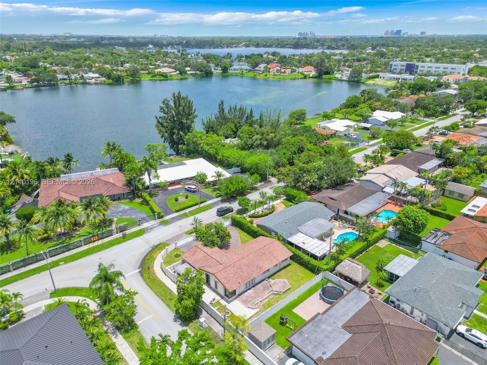 Aerial perspective of a lakefront residential lot featuring a tile roof, paved driveway, masonry wall perimeter, and mature landscaping