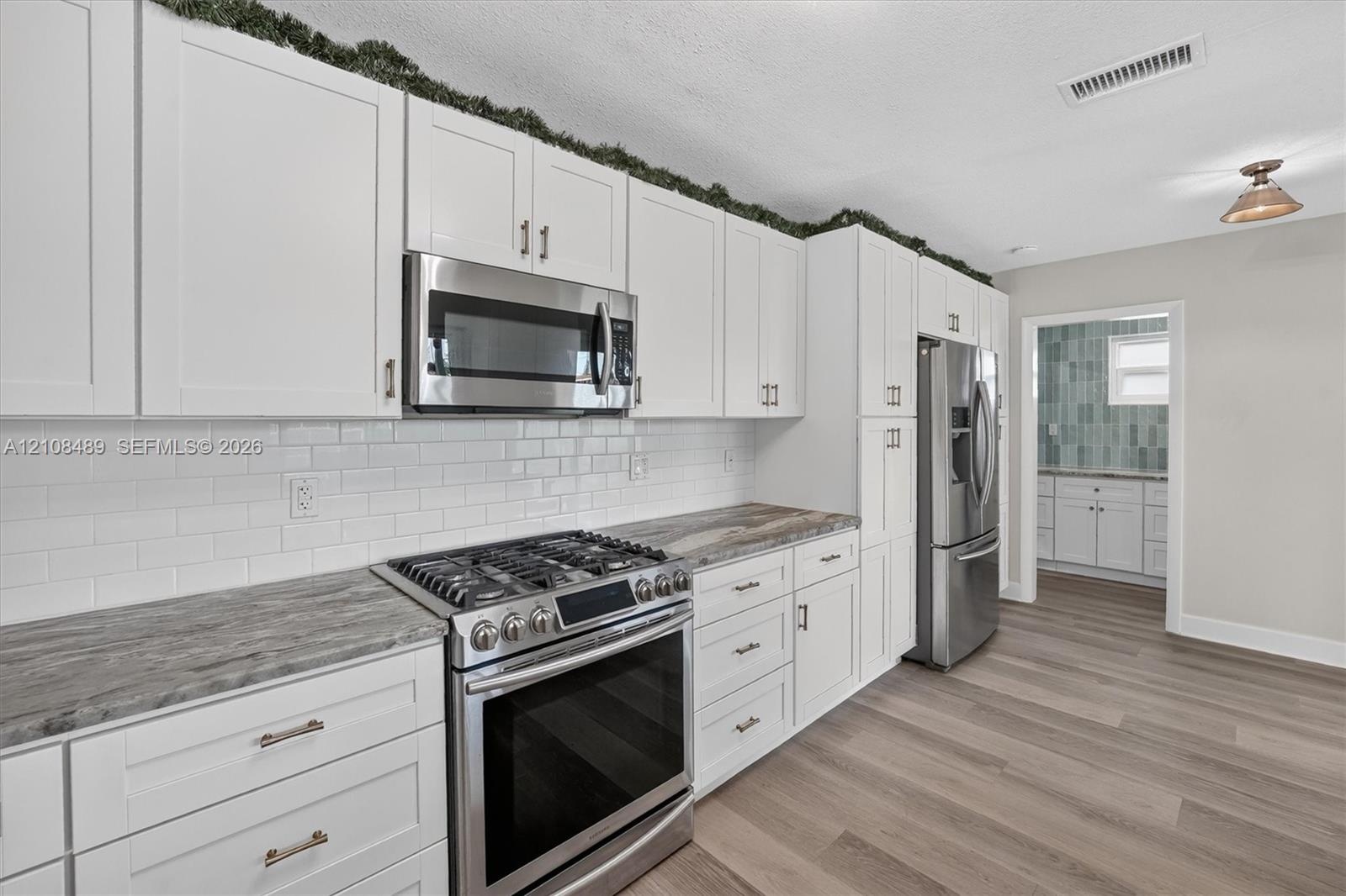 Kitchen featuring white shaker-style cabinetry, stone-patterned countertops, a white subway tile backsplash, stainless steel appliances, and wood-fini