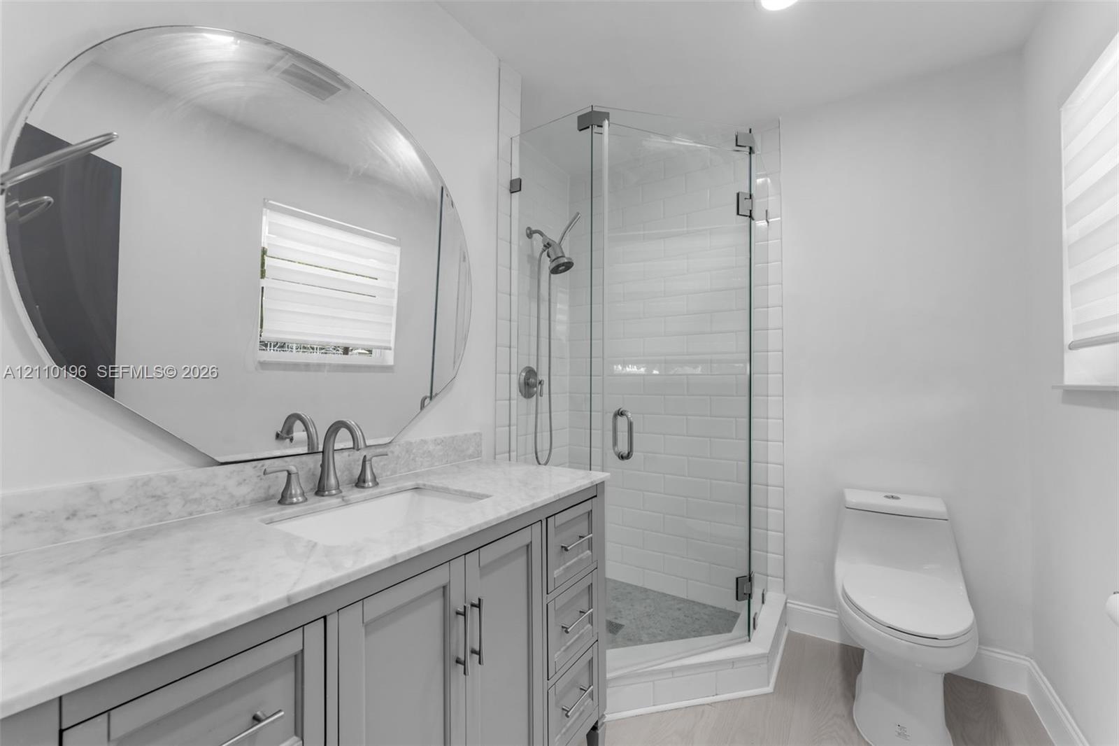 Bathroom featuring a gray vanity with a neutral stone countertop, an oversized circular mirror, a glass-enclosed shower with subway tile surround, and