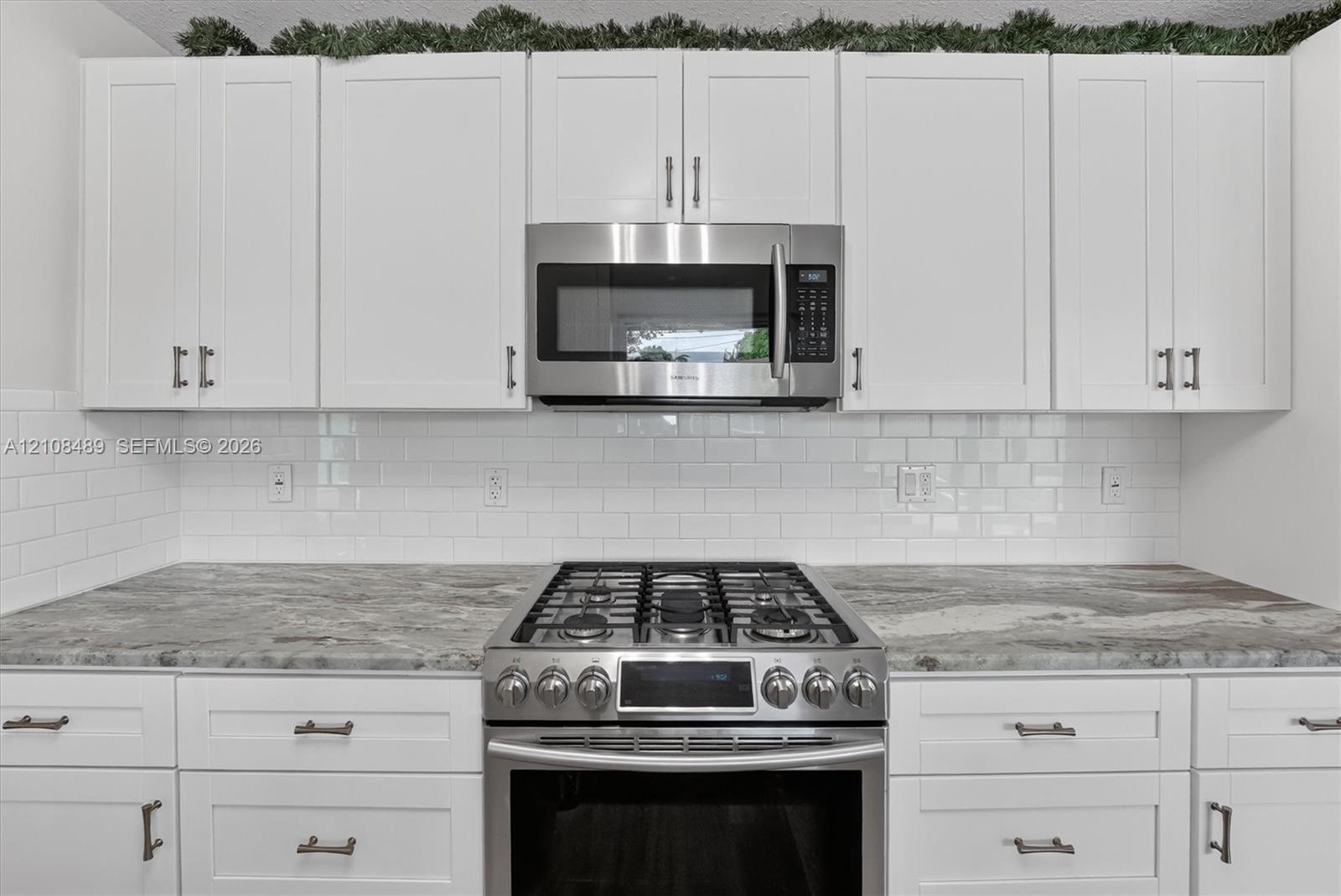 White shaker-style cabinetry with metallic hardware, stone-finish countertops, subway tile backsplash, and stainless steel gas range and built-in micr