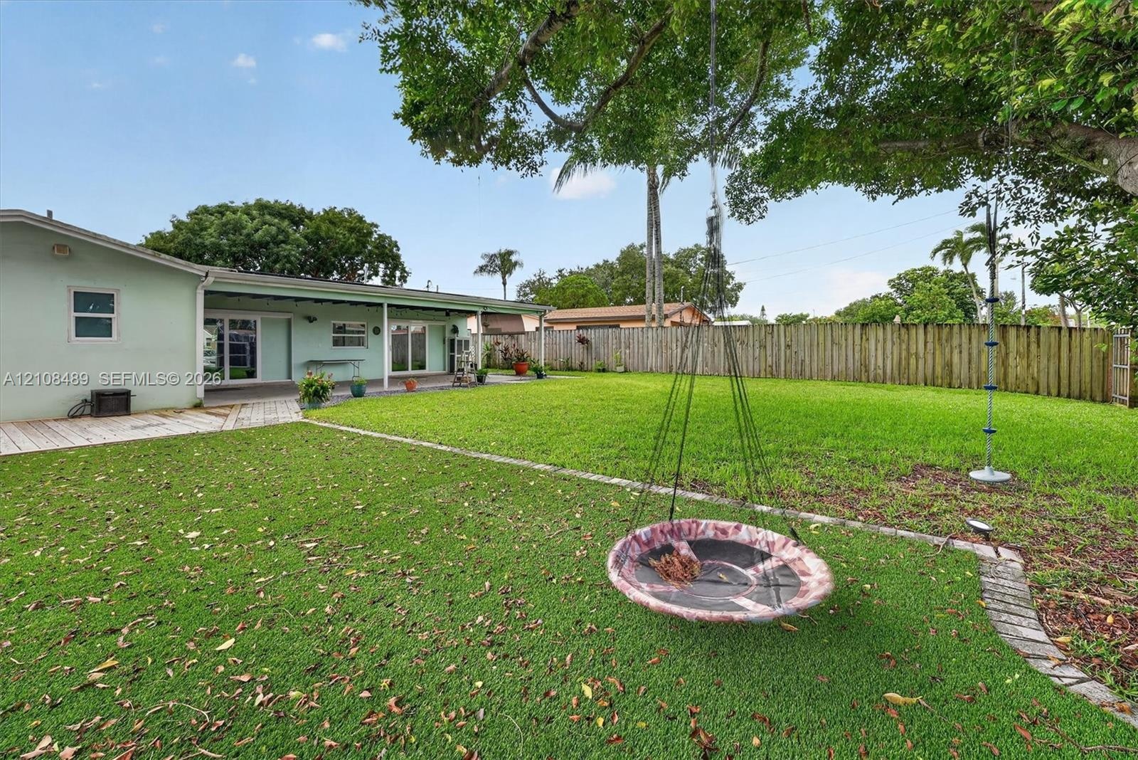 Expansive backyard featuring a covered patio, a manicured lawn, perimeter wood fencing, and mature trees