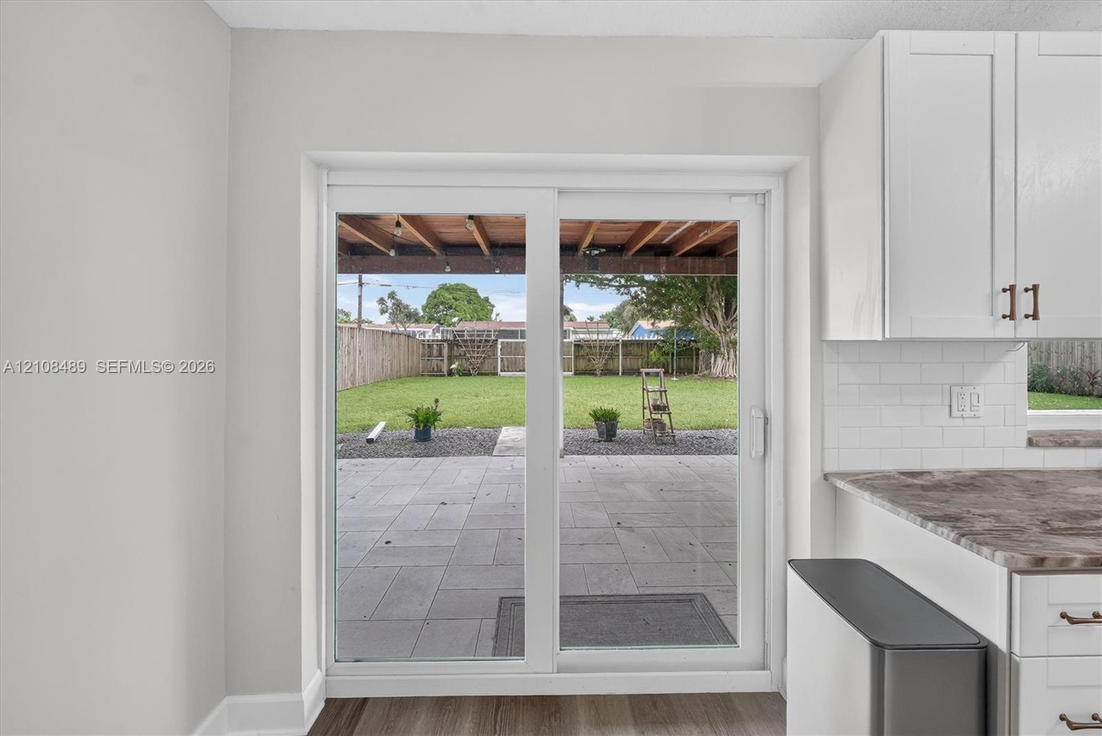 Sliding glass door opening to a concrete patio, covered pergola, fenced backyard with turf, and white shaker-style cabinetry with subway tile backspla