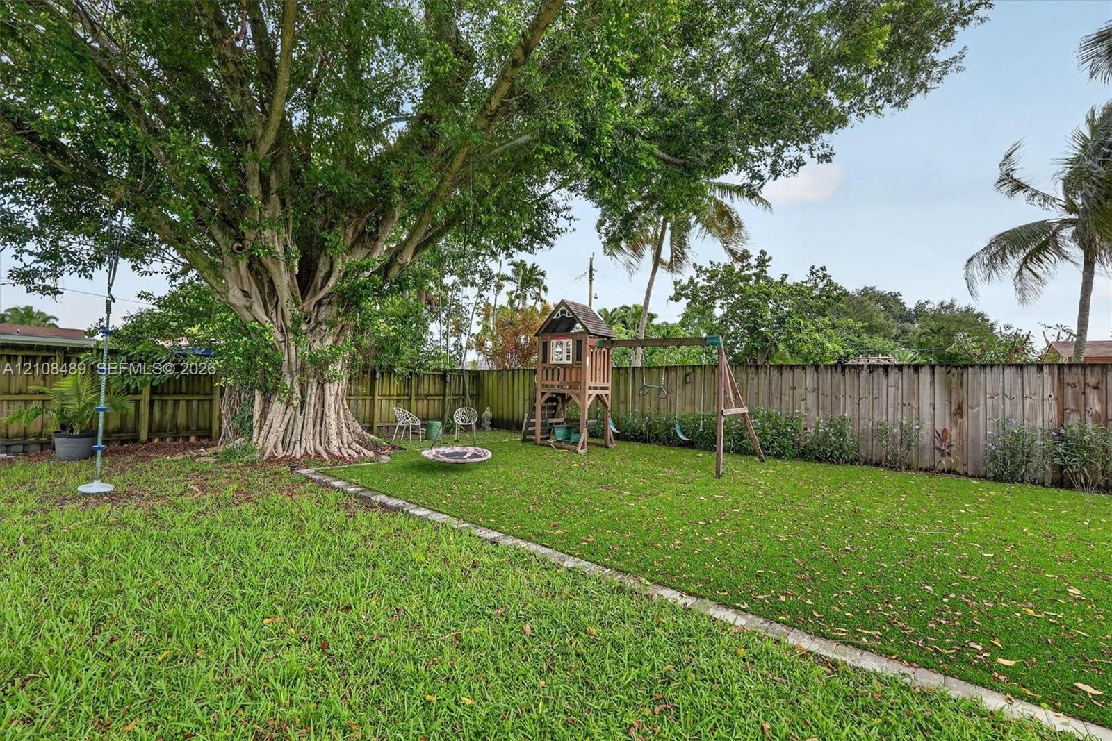 Backyard featuring synthetic turf, concrete paver edging, wood perimeter fencing, and a mature tree