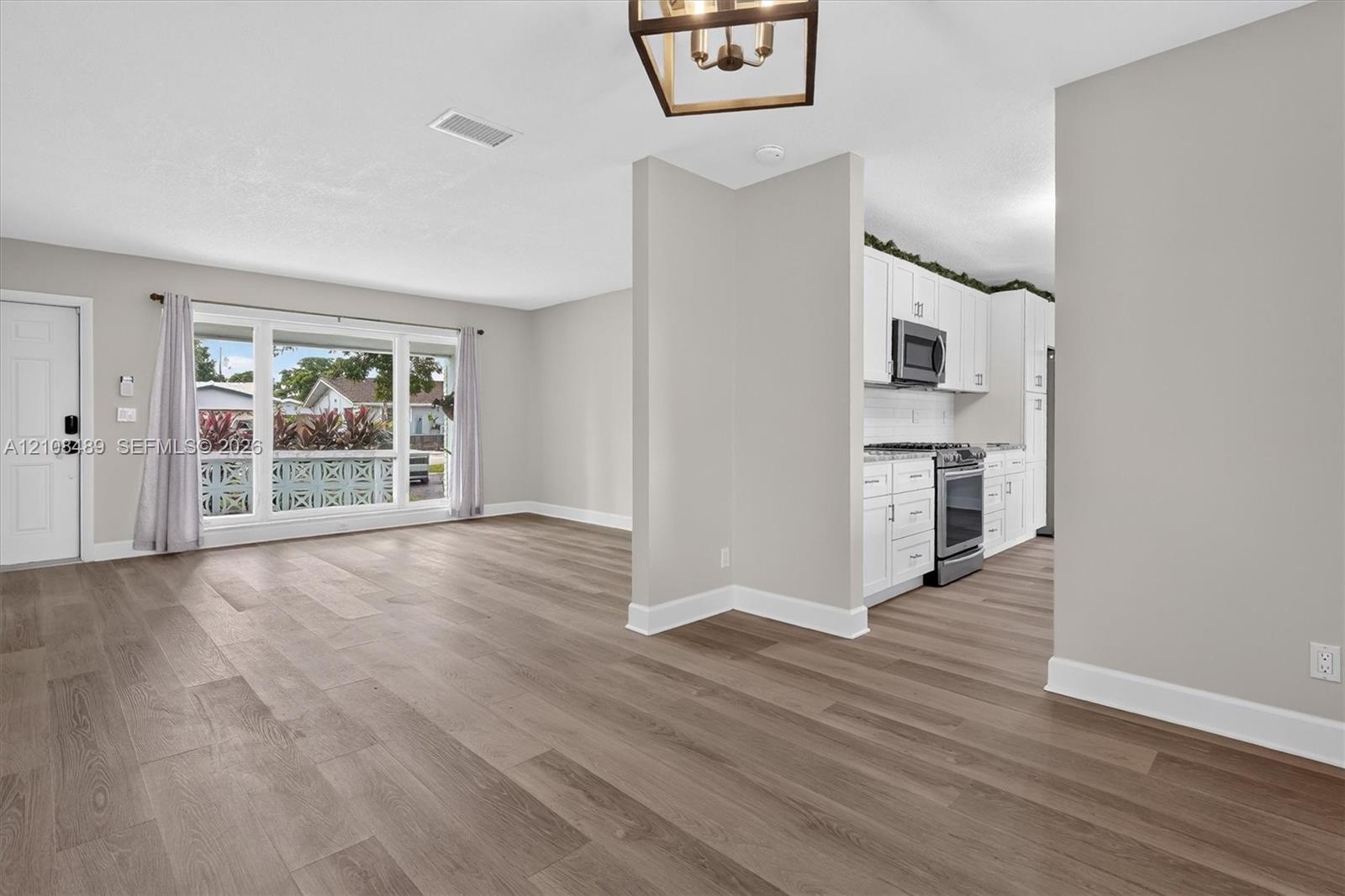 Open interior featuring wood-finish flooring, white cabinetry, integrated stainless steel appliances, a picture window, and a geometric pendant light 