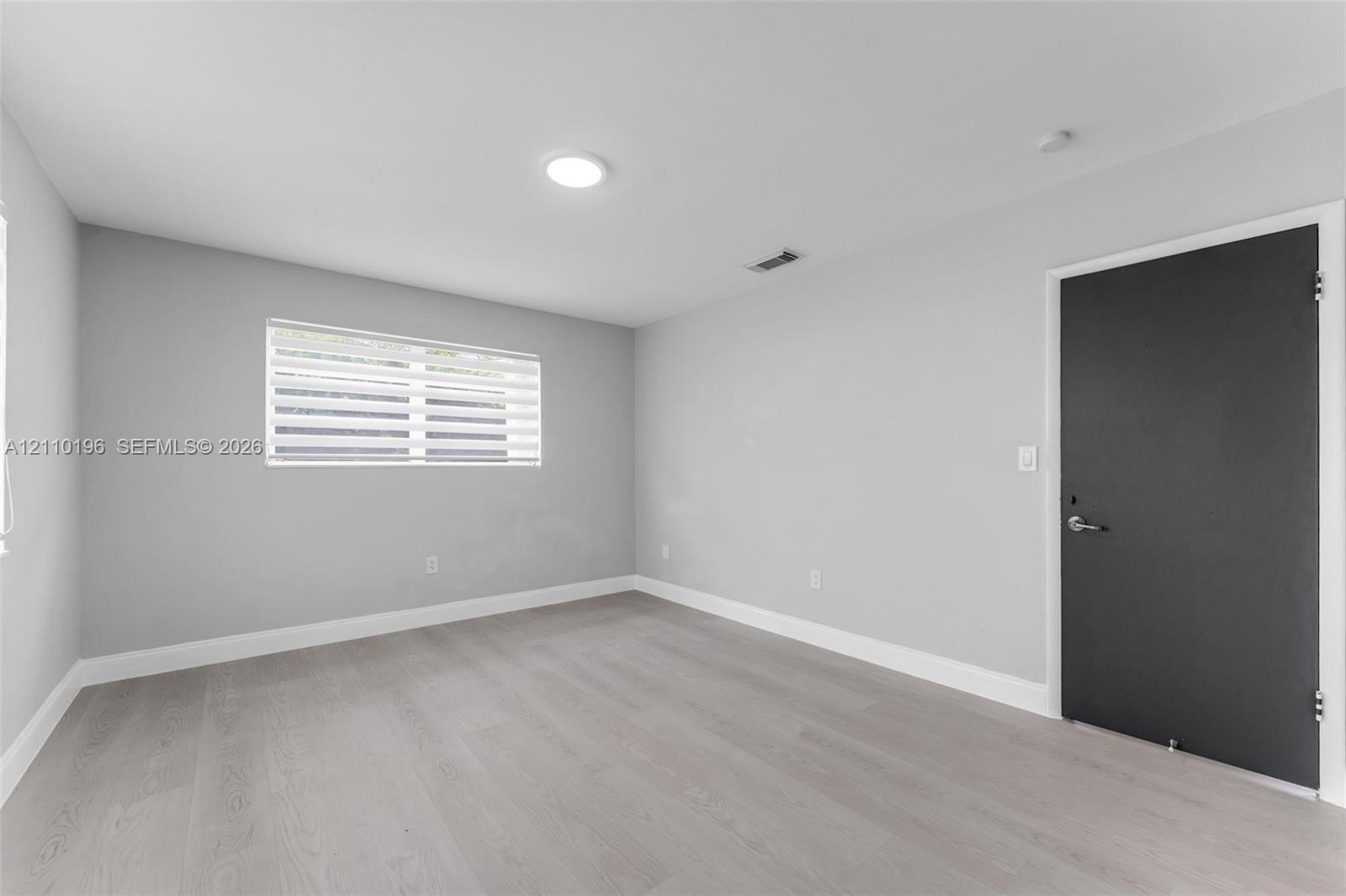 Neutral-toned interior space featuring wood-finish flooring, light gray walls, and a contrasting dark-finished door with metallic hardware