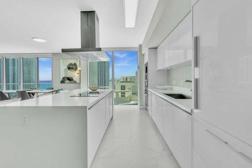 Sleek kitchen featuring an oversized island, integrated cooktop, stainless steel range hood, wall ovens, and custom white cabinetry