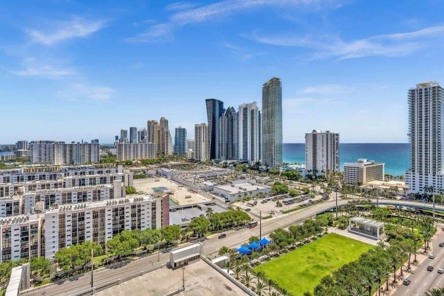 Expansive urban landscape featuring high-rise buildings, distant ocean, and a green space with mature trees