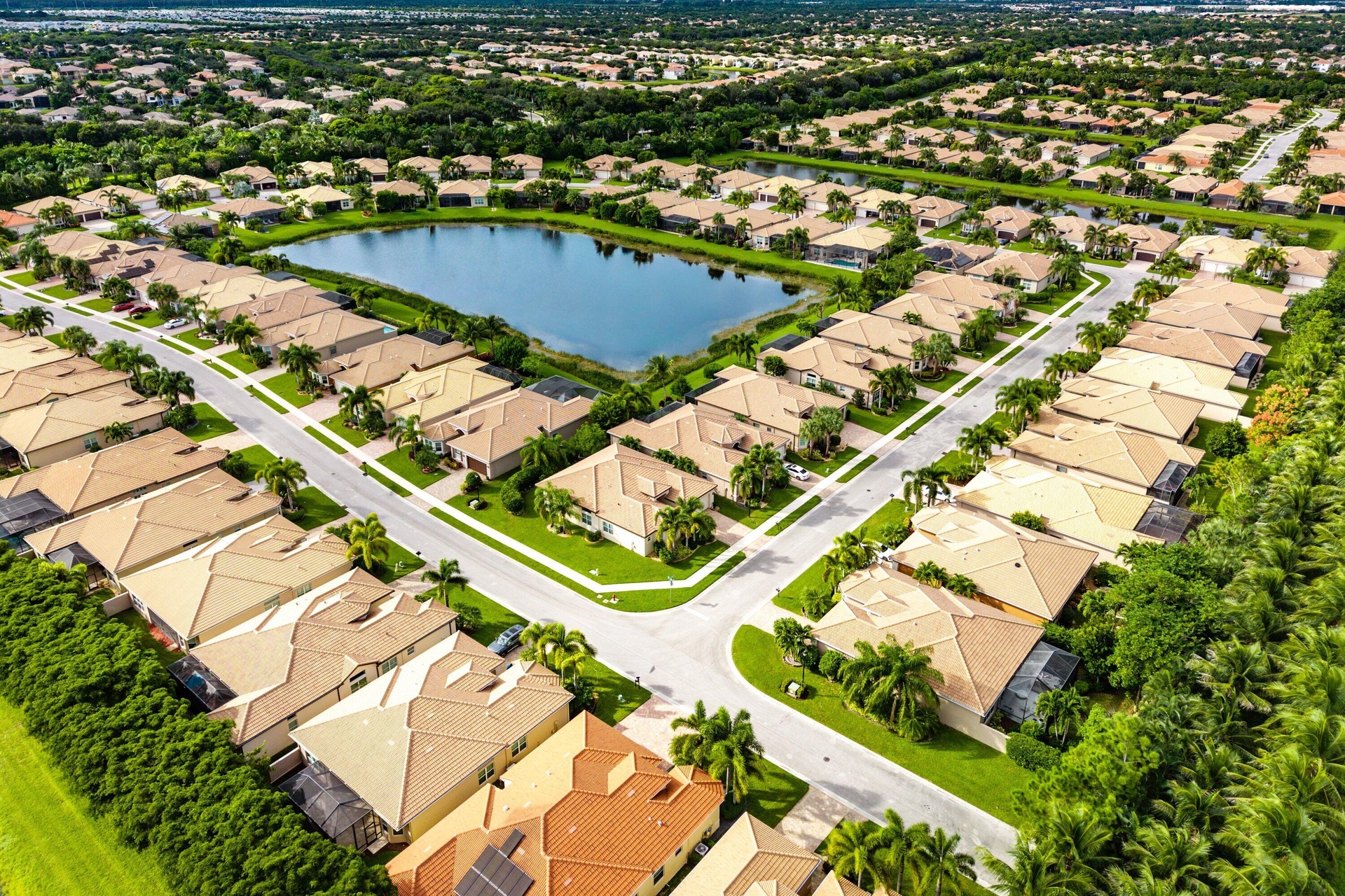 Aerial perspective of a planned community featuring a central lake, paved street networks, tile-roof residential structures, and mature palm trees