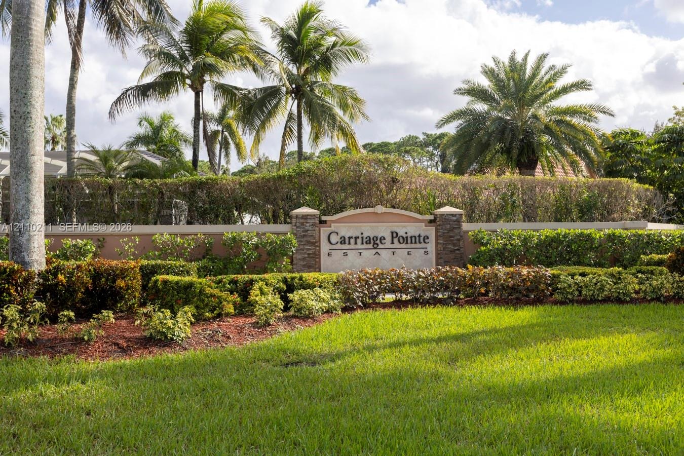Community entrance monument featuring stacked stone pillars, stucco masonry, and extensive tropical landscaping including manicured lawn and mature pa