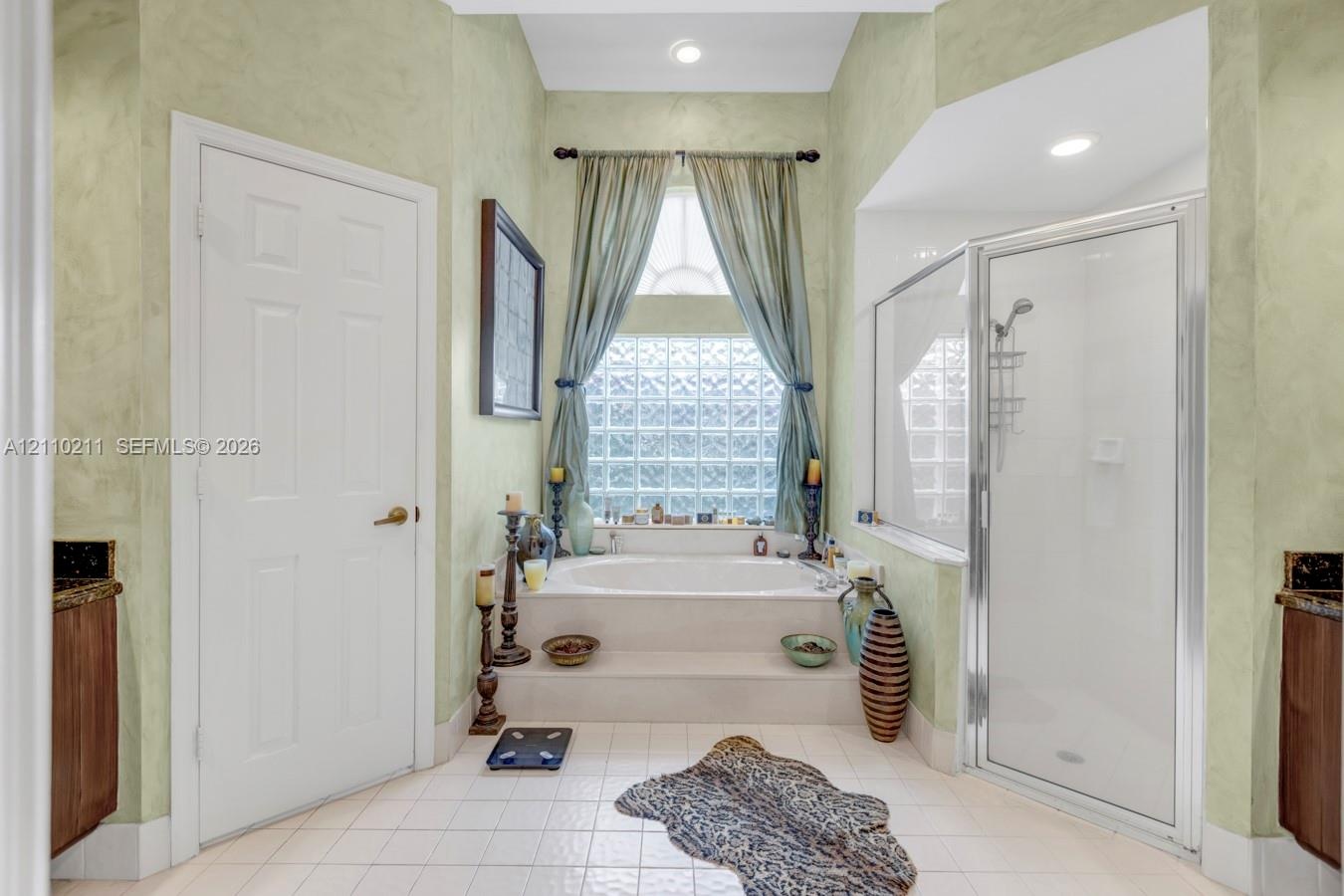 Bathroom featuring a recessed soaking tub with a tiled surround, glass block window, separate shower stall with a glass enclosure, and dual cabinetry 