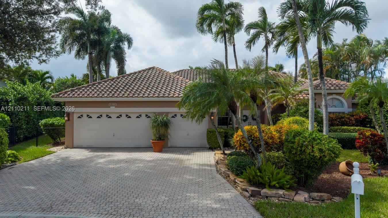 Single-story exterior featuring a clay tile roof, two-car garage with arched windows, and a paver stone driveway surrounded by mature palms and profes