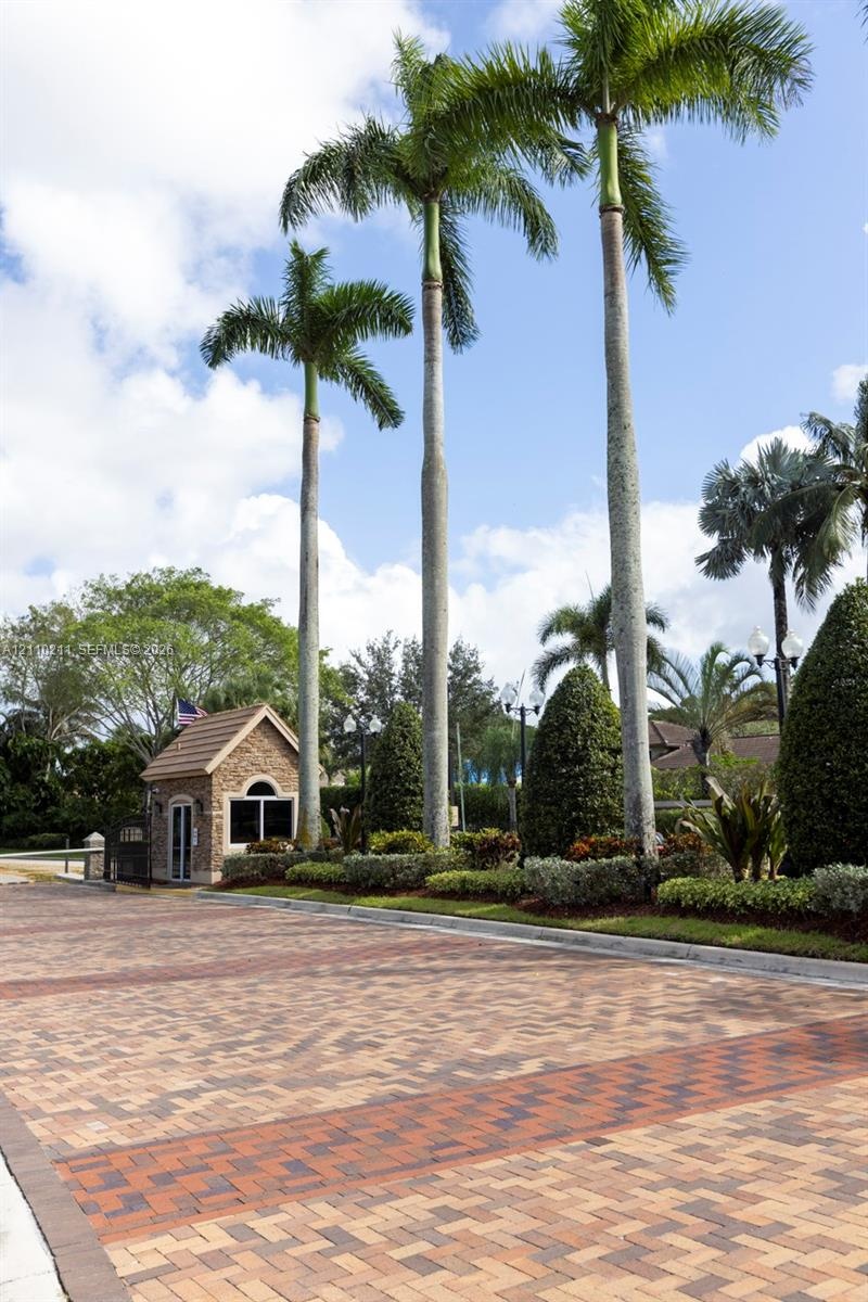 Gated entrance featuring a stone-clad guardhouse, intricate paver driveway, manicured landscaping, and mature palm trees