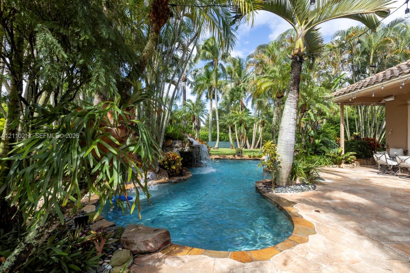 Freeform swimming pool with a stone-accented waterfall, natural stone patio, and mature tropical landscaping