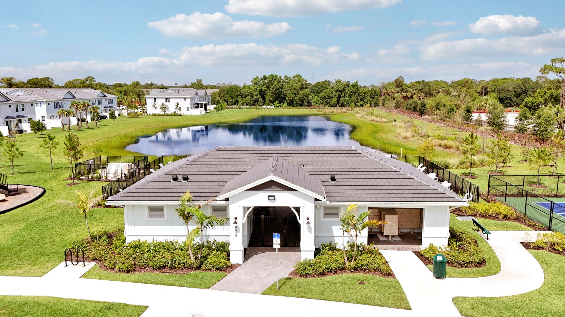 Community clubhouse featuring a dark tile roof, exterior lighting fixtures, and a paved walkway, situated on grounds with a retention pond and sport c