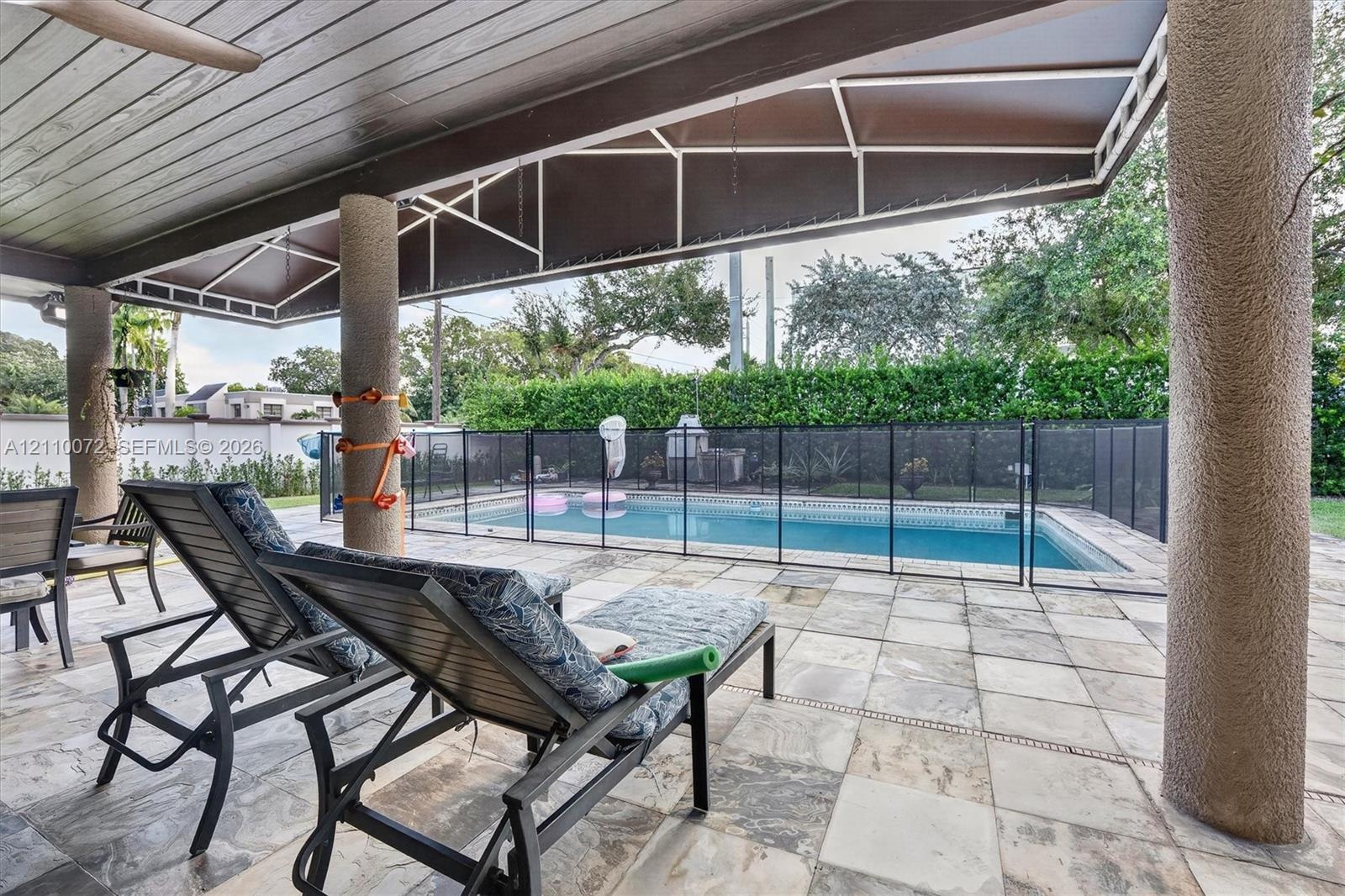 Covered patio with a wood-plank ceiling, structural support columns, and stone tile flooring, featuring an inground swimming pool with a black mesh sa