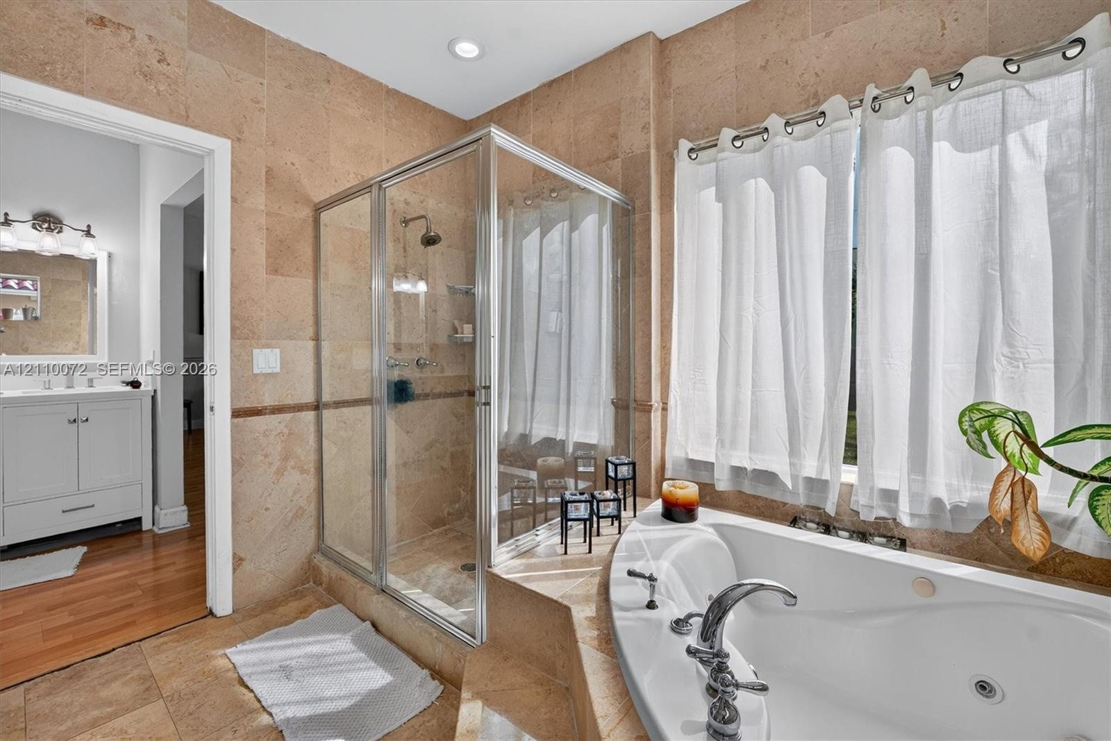 Full-sized jetted soaking tub with a tiled surround, glass-enclosed shower, natural stone wall and floor tile, recessed lighting, and a white vanity w