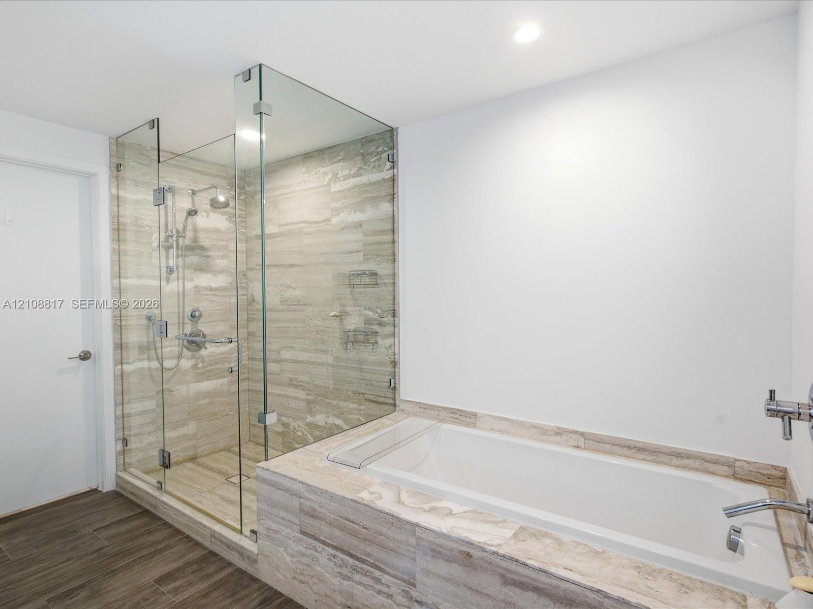 Walk-in glass-enclosed shower with a rainfall showerhead, alongside a built-in soaking tub featuring stone-finish tile surround and wood-finish floori