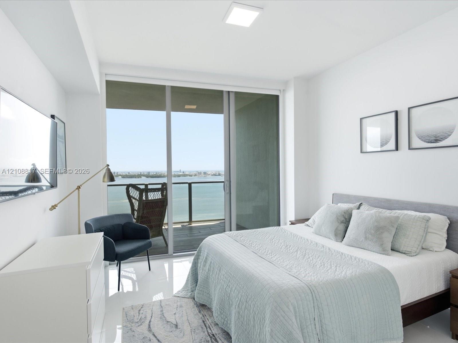 Floor-to-ceiling glass sliding doors opening to a private balcony, polished tile flooring, and prominent water and city skyline backdrop