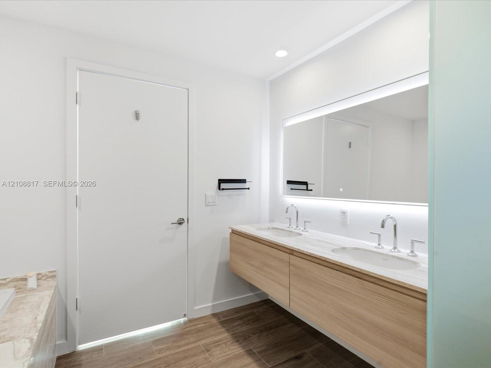 Floating dual-sink vanity with light wood cabinetry, stone-surface countertop, integrated LED mirror lighting, and wood-finish flooring