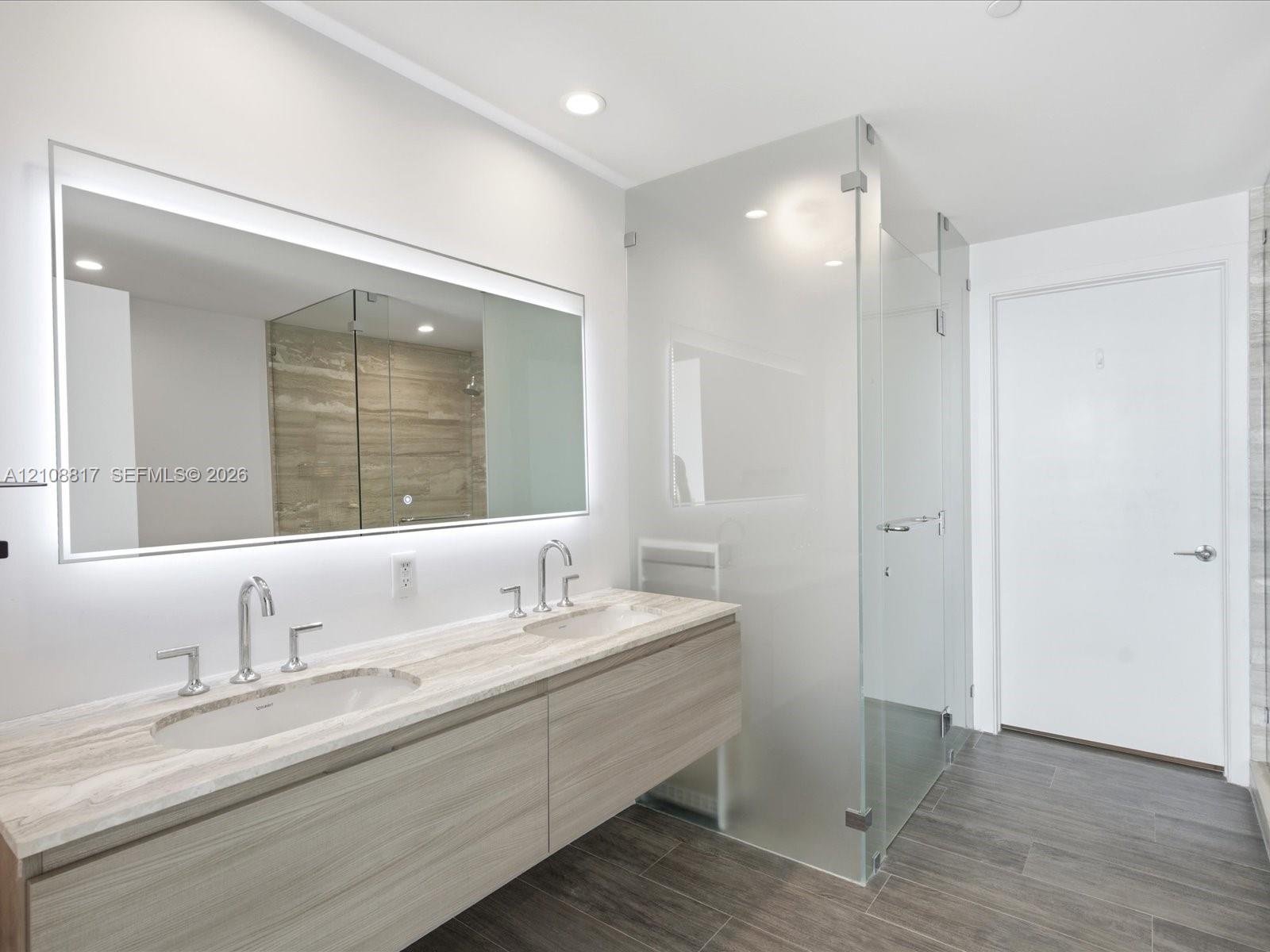 Floating dual-sink vanity featuring stone countertops and polished chrome widespread faucets, complemented by a large back-lit mirror, glass-enclosed 
