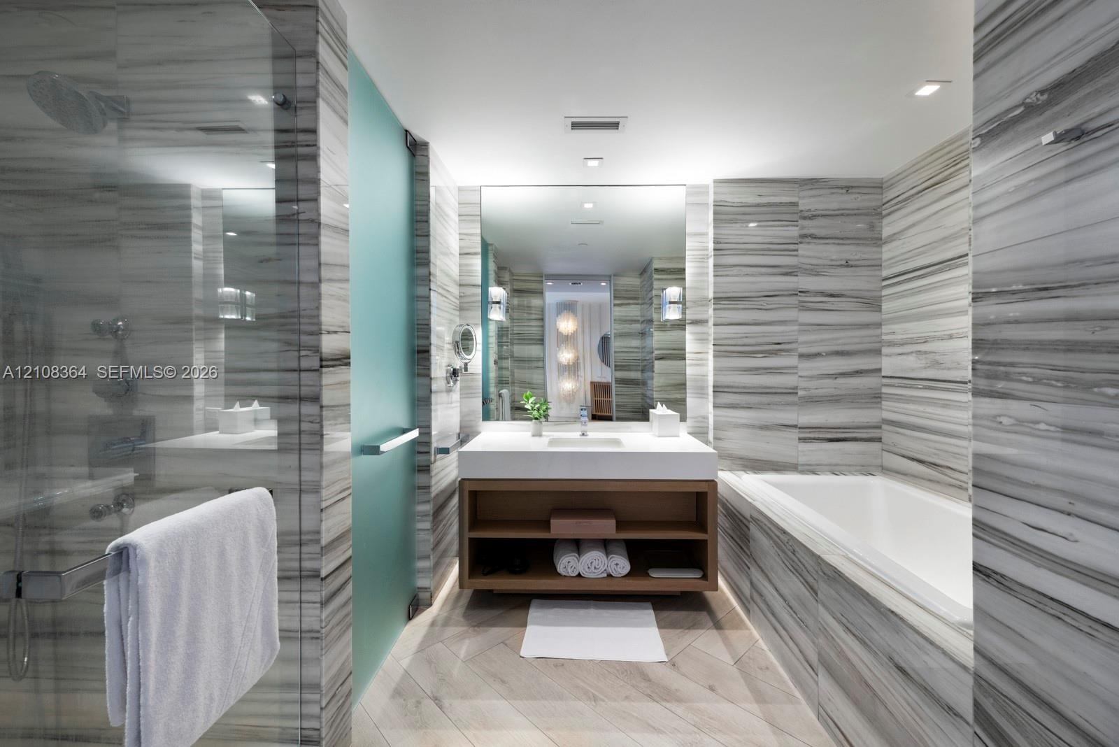 Bathroom featuring floor-to-ceiling stone tile, a soaking tub, a glass-enclosed shower, a floating wood-finish vanity, and recessed lighting