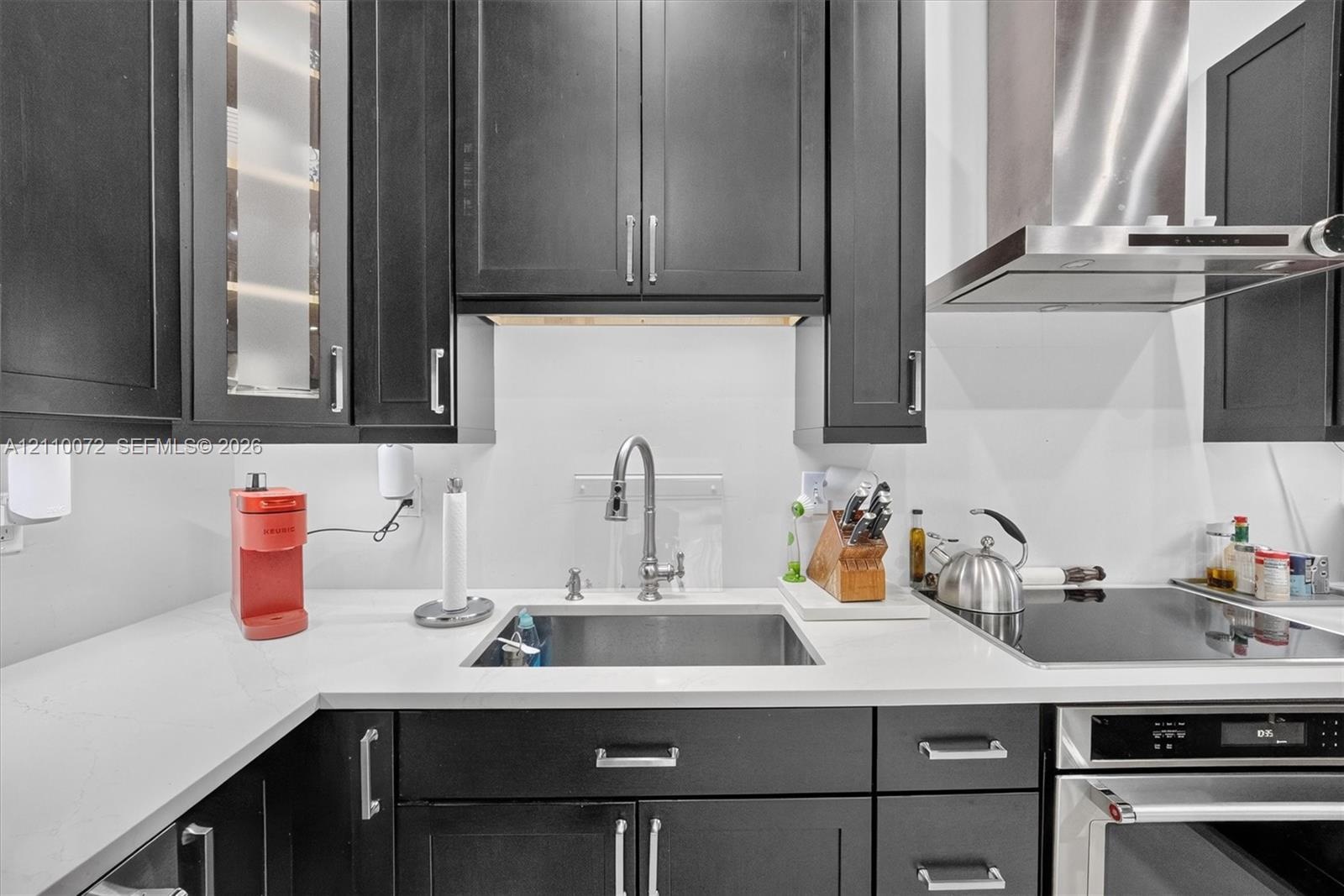 Dark finish cabinetry with glass-front accents, solid-surface countertops, stainless steel undermount sink, gooseneck faucet, and integrated stainless