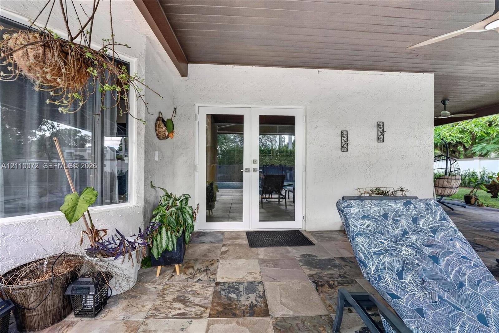 Covered patio featuring multi-tonal stone flooring, white textured exterior wall, dual-pane glass French doors, and a wood-finish ceiling with an inte