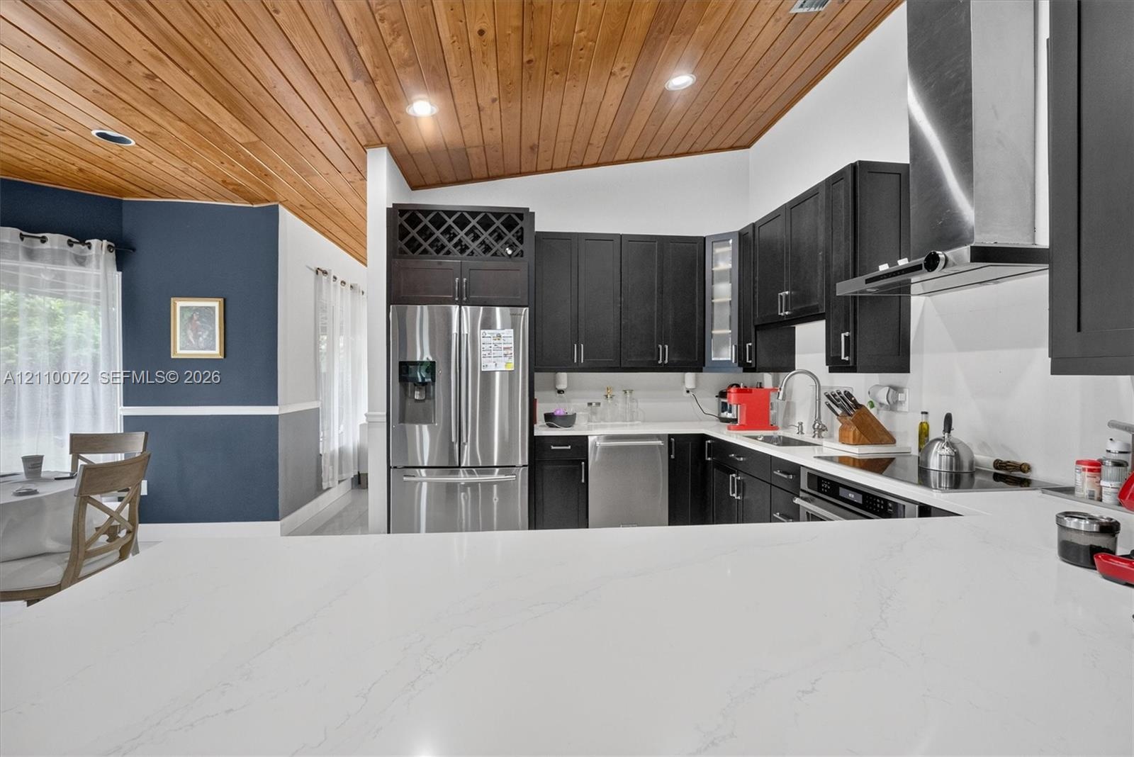 Kitchen featuring vaulted wood-paneled ceiling, white stone-finish countertops, stainless steel appliances, dark wood cabinetry with built-in wine sto
