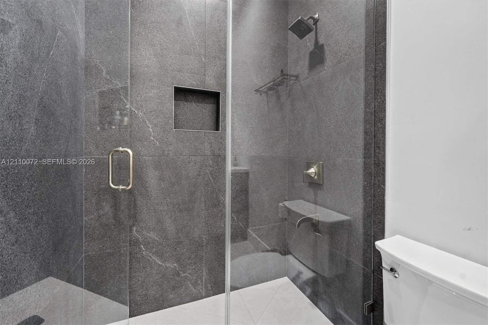 Walk-in shower featuring floor-to-ceiling stone-finish tile, frameless glass door, built-in wall niche, brushed metallic fixtures, and a stationary sh