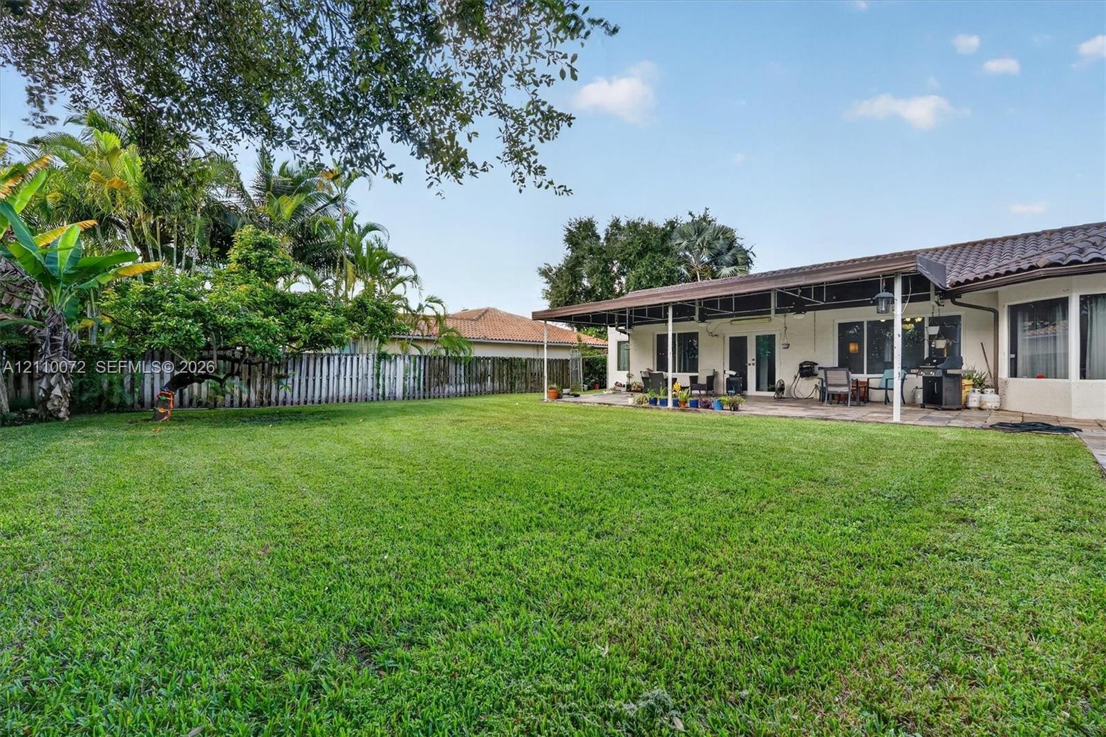Expansive backyard featuring a manicured lawn, mature trees, a perimeter privacy fence, and a covered patio with a tile roof