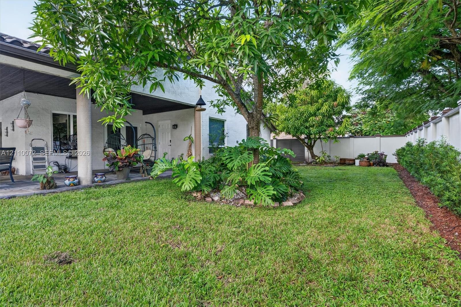 Covered concrete patio featuring structural columns, expansive lawn, stone-bordered landscaping, and perimeter privacy wall