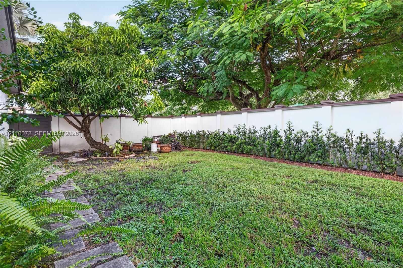 Private backyard featuring a manicured lawn, stone paver pathway, mature trees, and a solid perimeter wall with hedge landscaping