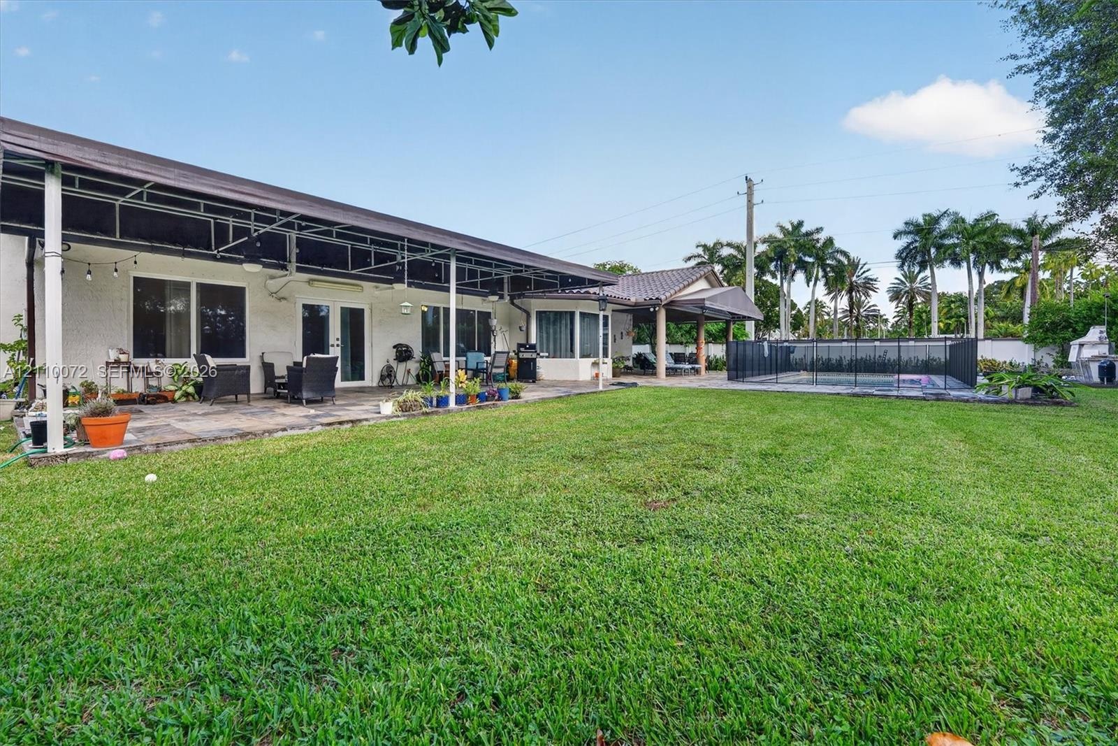 Backyard featuring an expansive lawn, stone-paved patio, covered terrace, in-ground swimming pool with safety fencing, and mature palm trees
