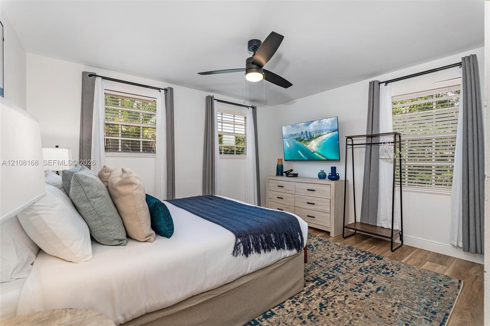 Bedroom featuring wood-finish flooring, three windows with plantation-style shutters, and a central ceiling fan with integrated lighting