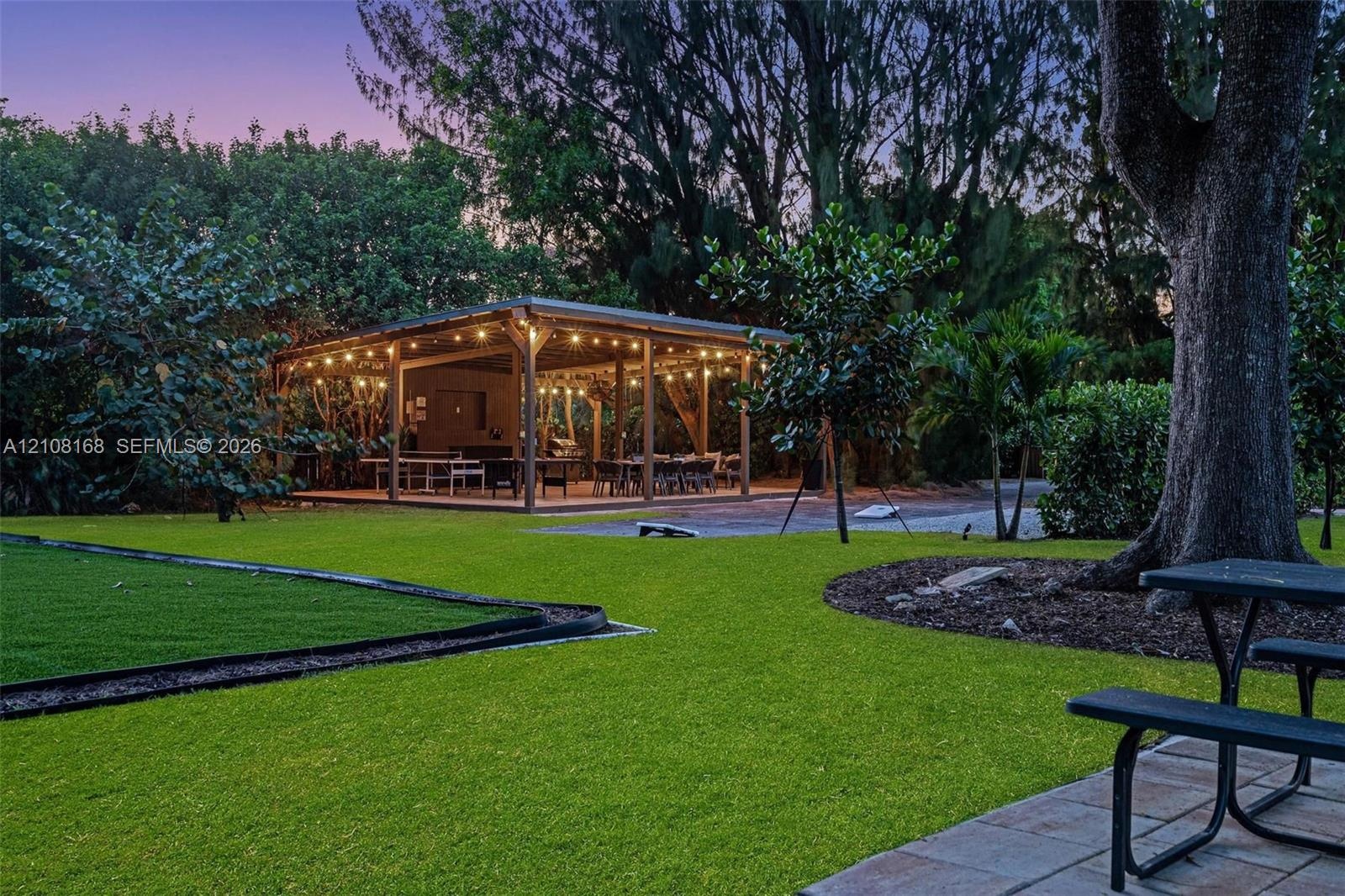 Expansive backyard featuring a covered wood-frame pavilion, manicured lawn, stone paver patio, and mature trees