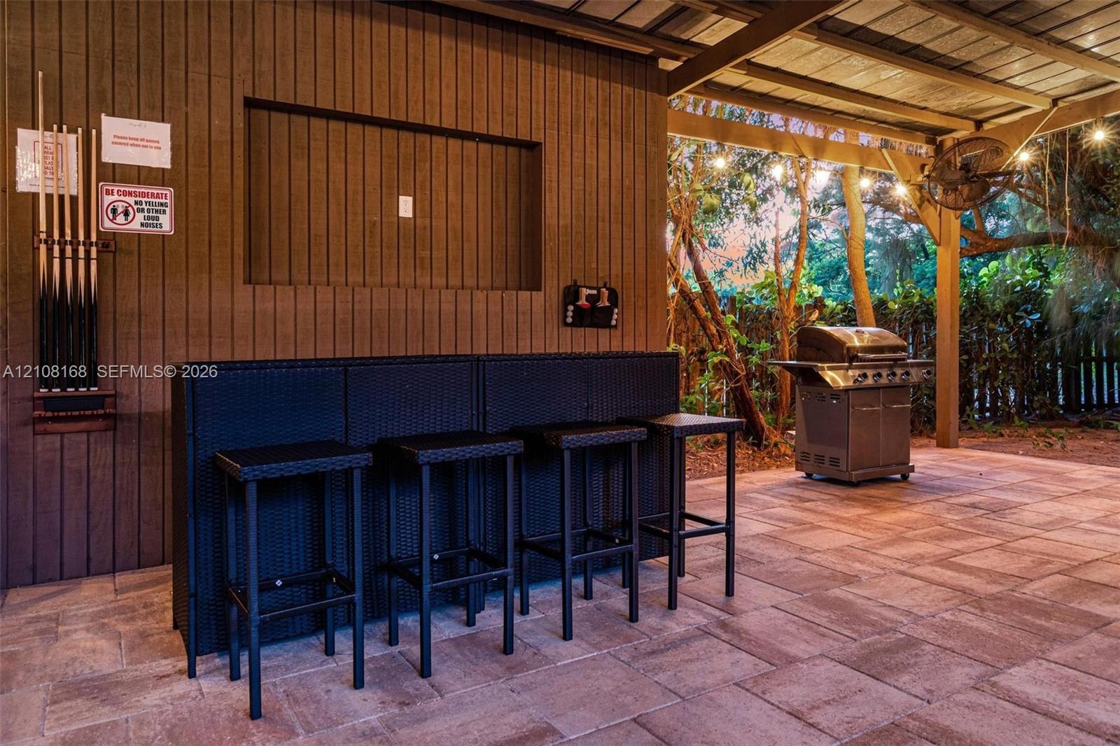 Covered outdoor patio featuring paver flooring, vertical wood-plank siding, built-in television niche, and overhead string lighting