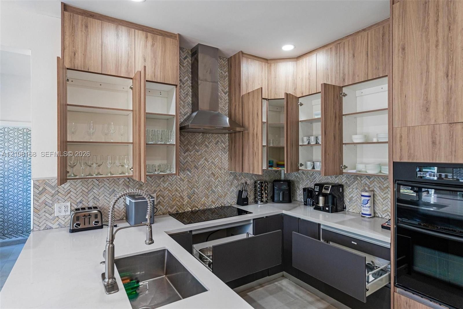 Kitchen featuring custom wood-finish cabinetry, stone-surface countertops, a herringbone-patterned tile backsplash, a stainless steel range hood, and 