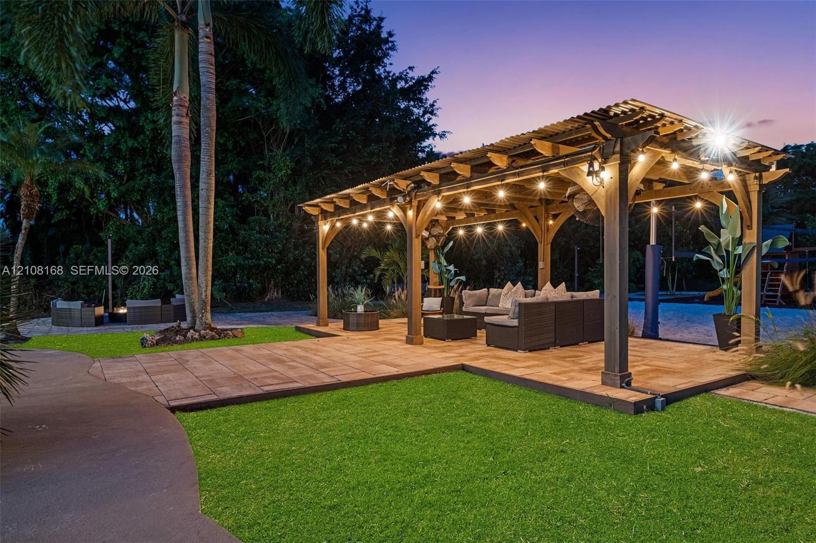 Backyard featuring a wood-frame pergola, stone-paver patio, manicured lawn, and mature palm trees