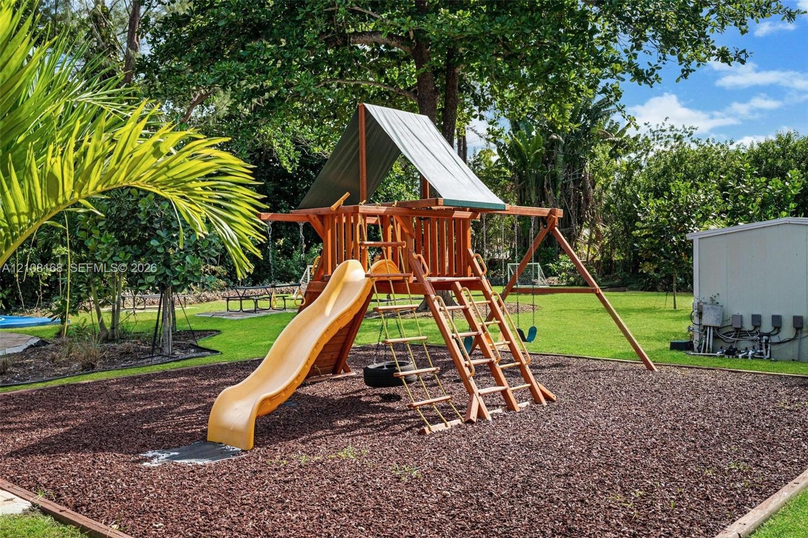 Expansive backyard featuring a wood-frame playground structure, rubberized ground cover, manicured lawn, and mature canopy trees