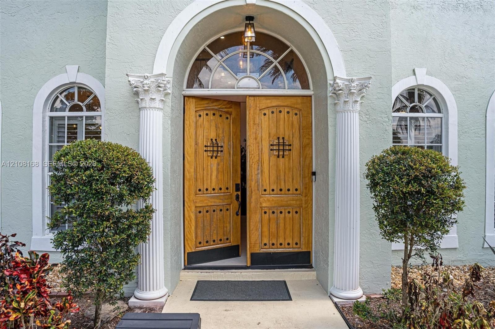 Entryway featuring dual wood-finish doors with decorative metal accents, an arched transom window, fluted columns with ornate capitals, and arched win