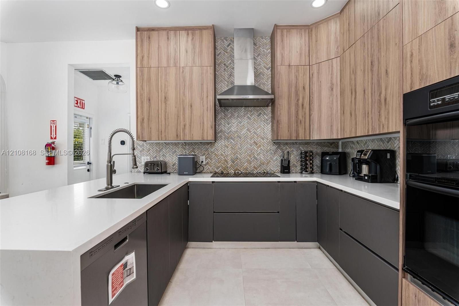 Kitchen featuring dual-tone cabinetry with wood-finish uppers, quartz countertops, herringbone-patterned tile backsplash, stainless steel range hood, 