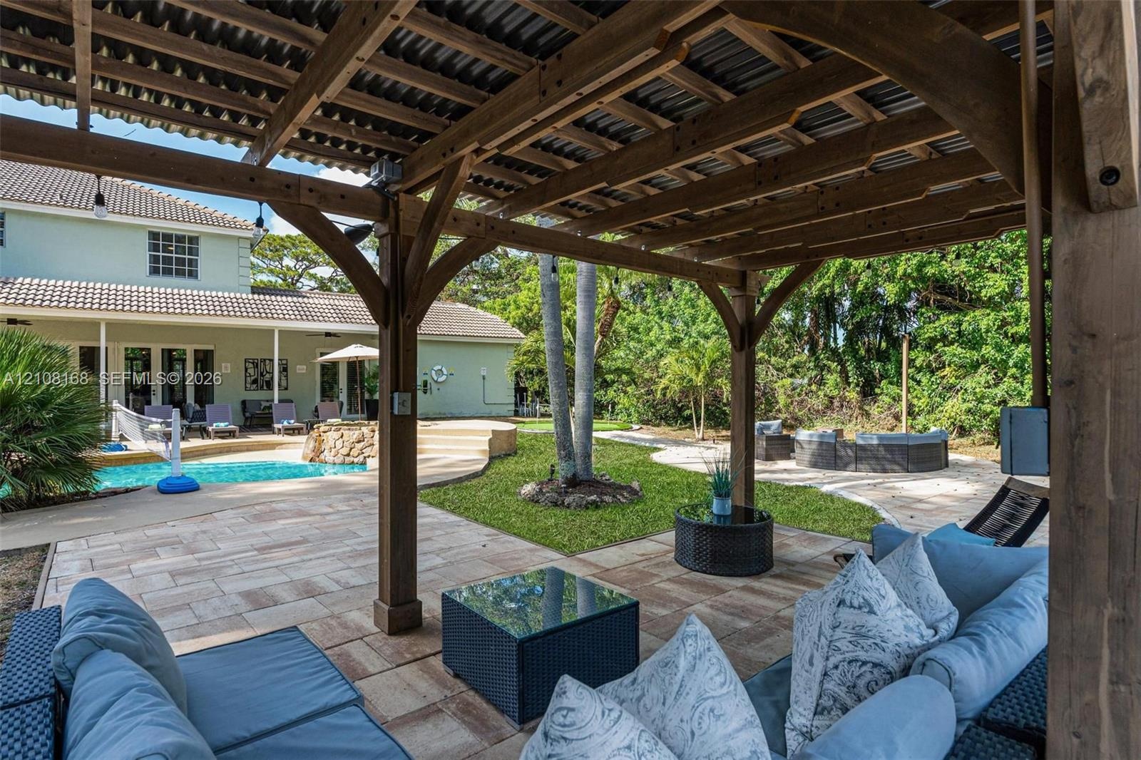 Covered wood-frame pergola with a corrugated roof, expansive stone-paver patio, swimming pool with a raised stone-faced spillway, and mature landscapi
