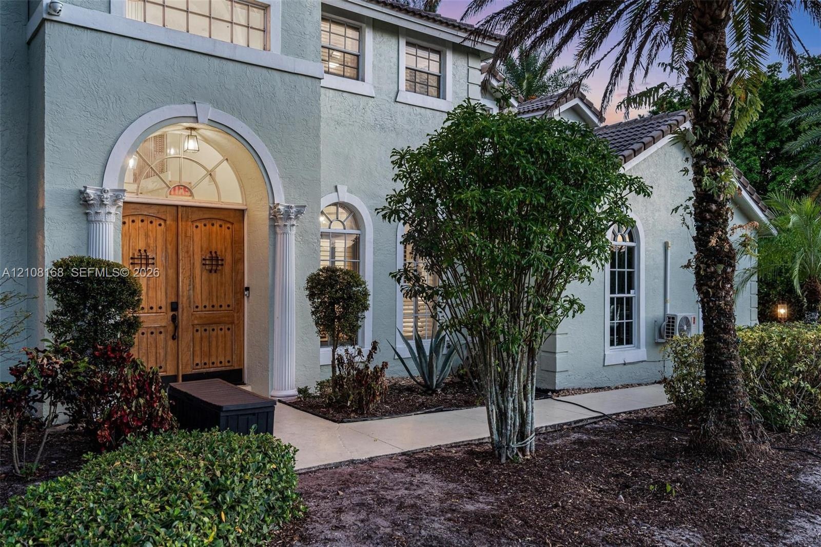 Stucco exterior featuring double arched wood front doors with decorative iron detailing, classical architectural columns, arched windows, and a paved 