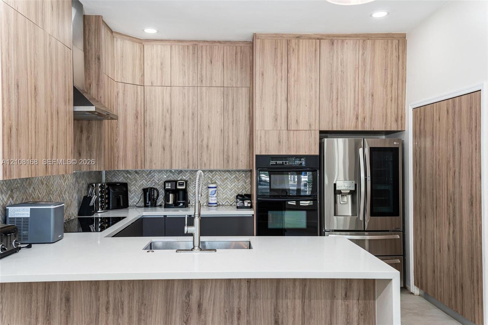 Kitchen featuring wood-finish cabinetry, white solid-surface countertops, a herringbone-patterned tile backsplash, a stainless steel undermount sink w