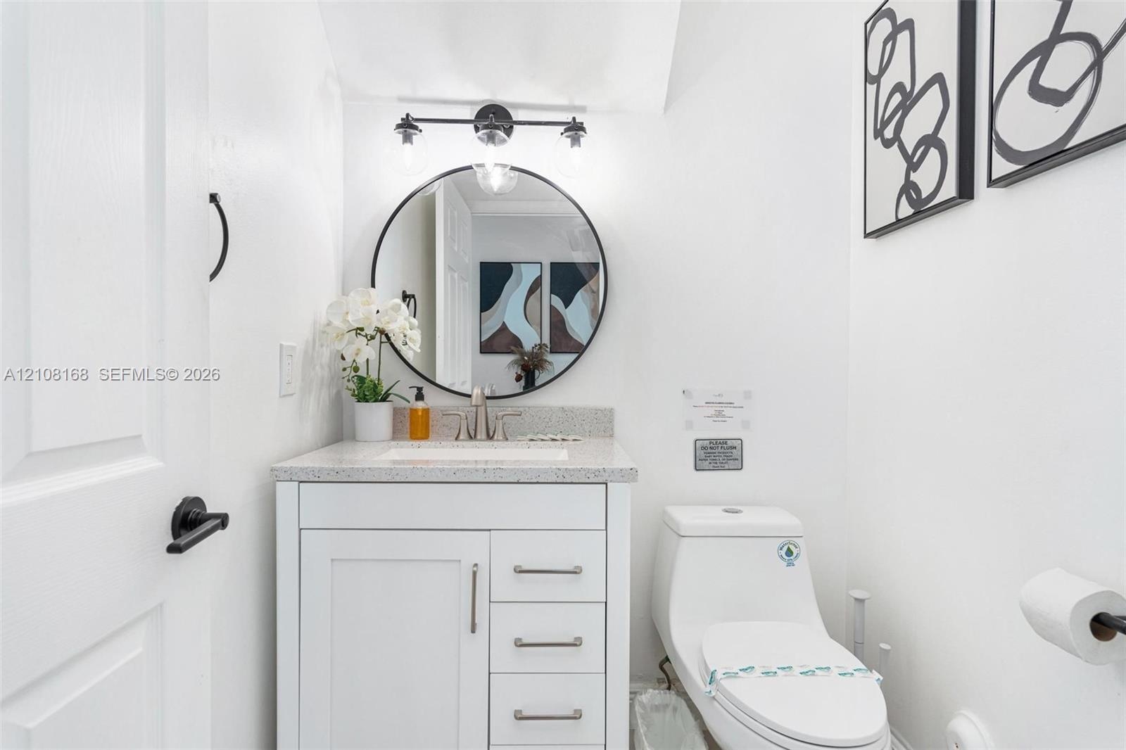 White vanity cabinet with a speckled countertop, integrated sink, circular wall-mounted mirror, three-light matte black fixture, and low-profile toile